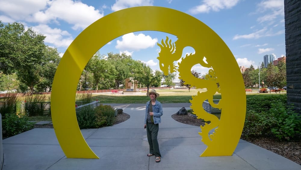 Audrey Bergner standing beneath a large yellow dragon sculpture in Calgary Chinatown, Alberta, Canada, with landscaped park space, trees and downtown buildings visible behind her.