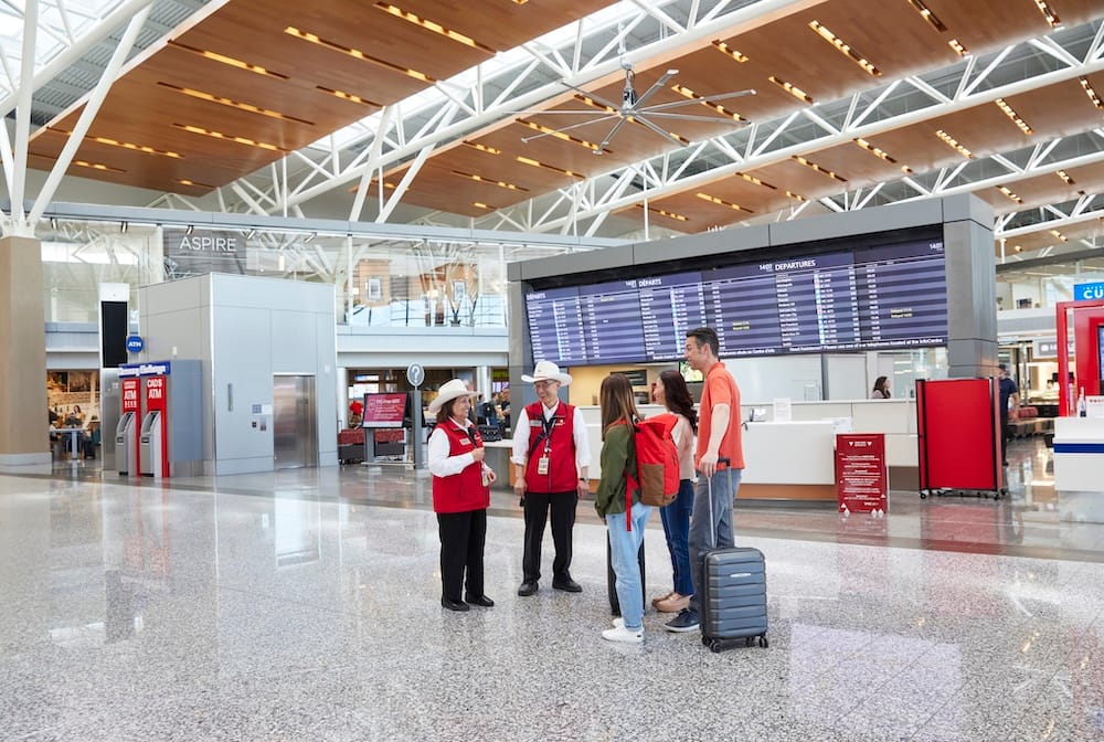 Viajeros con valijas de ruedas conversando con personal del aeropuerto debajo del tablero de salidas dentro de Calgary International Airport, YYC, en Calgary, Alberta, Canadá.