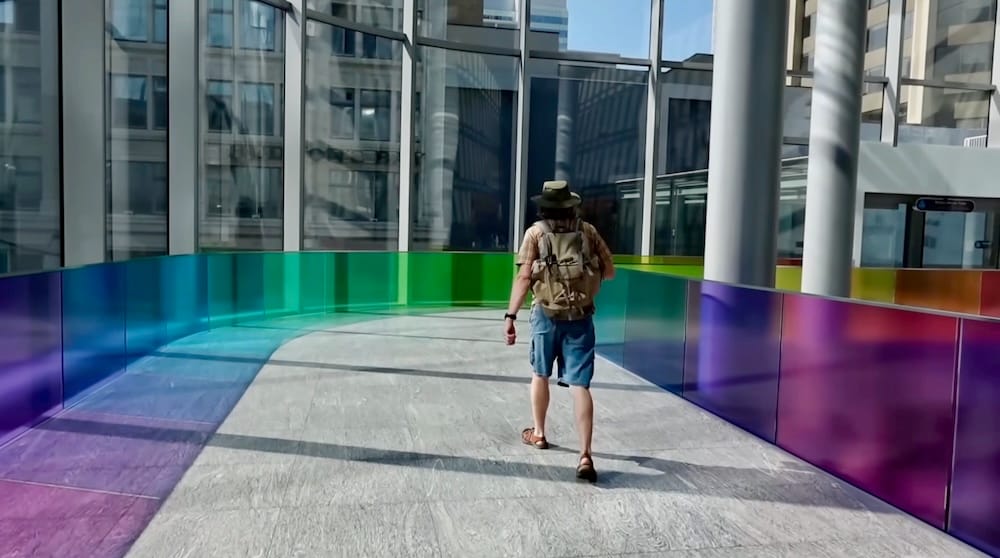 Samuel Jeffery caminando por la colorida red +15 Skywalk de Calgary, Alberta, Canadá Samuel Jeffery camina por la red +15 Skywalk de Calgary, Alberta, Canadá, junto a paneles de vidrio de colores y grandes ventanales con edificios del centro al fondo, llevando una mochila grande.