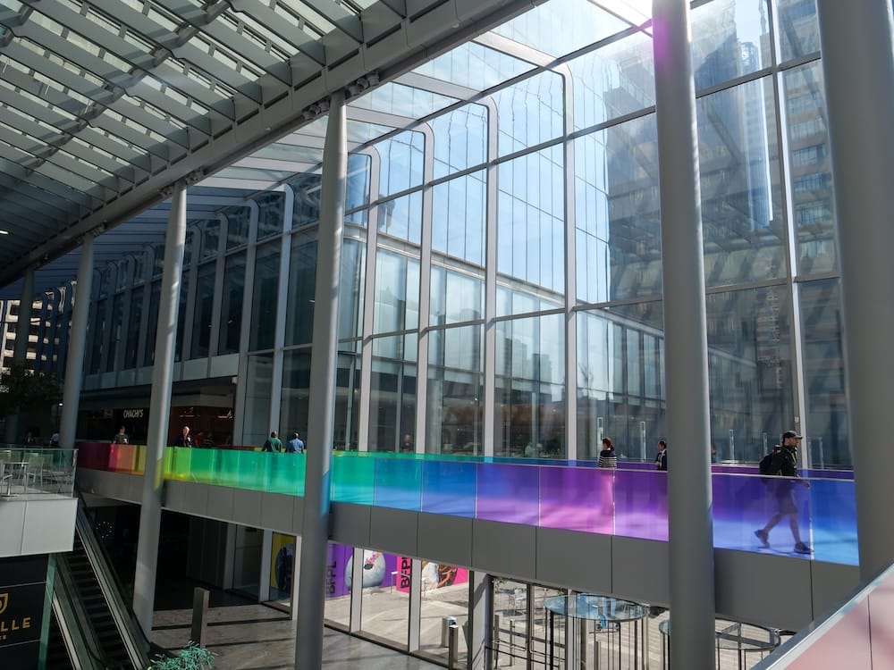 Calgary +15 Skywalk con pasarela de vidrio y paneles de colores en el centro de Calgary, Alberta, Canadá La red +15 Skywalk de Calgary, Alberta, Canadá, muestra una pasarela elevada con techo de vidrio, paneles de colores iluminados y torres de oficinas del centro visibles a través de los grandes ventanales.
