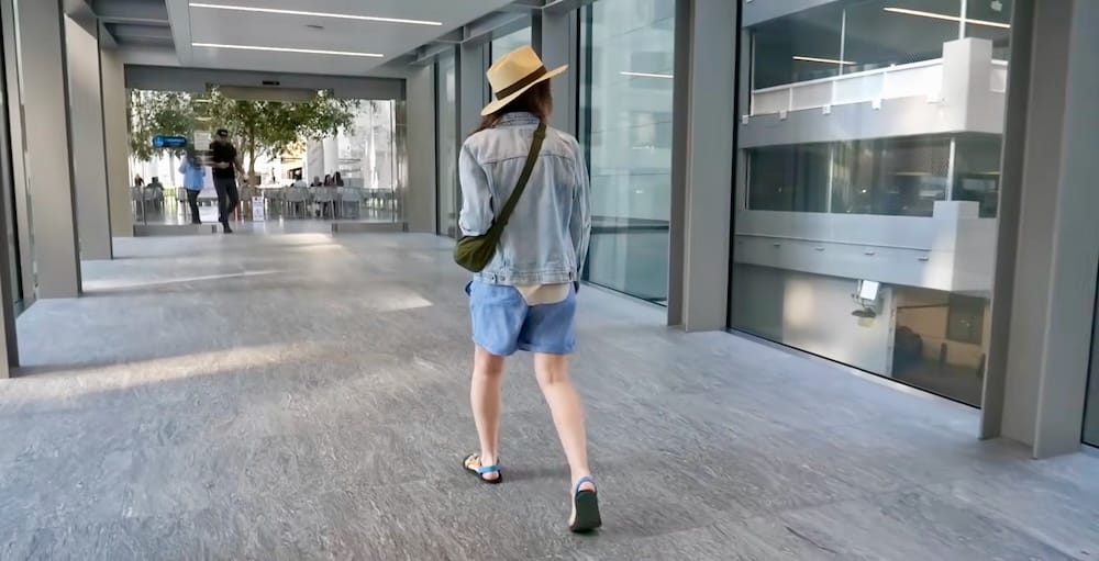 Audrey Bergner caminando por una sección vidriada del Calgary +15 Skywalk en el centro de Calgary, Alberta, Canadá, con el corredor peatonal elevado continuando hacia un área pública interior más concurrida.