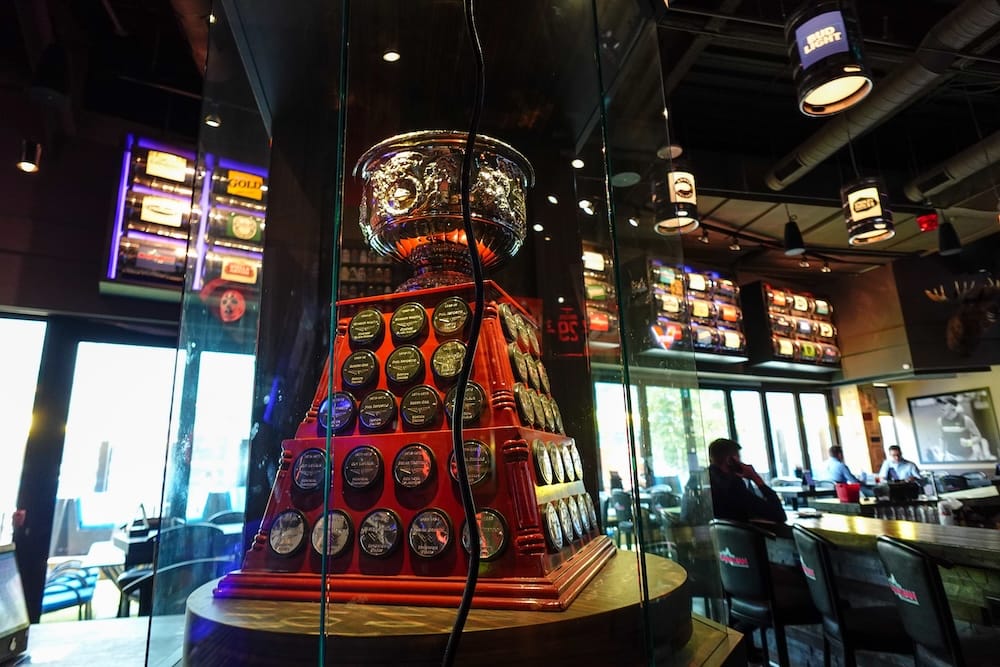Exhibición de un trofeo de hockey dentro de The Canadian Icehouse en Edmonton, Alberta, protegido por una vitrina de vidrio y rodeado de recuerdos deportivos, asientos de barra y paneles iluminados.