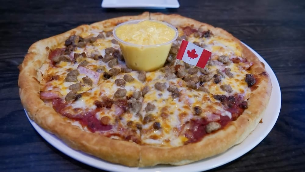 Primer plano de la pizza Meatza en The Canadian Icehouse de Edmonton, Alberta, con borde grueso, queso derretido, varias carnes, un recipiente de salsa de queso y una pequeña bandera canadiense encima.
