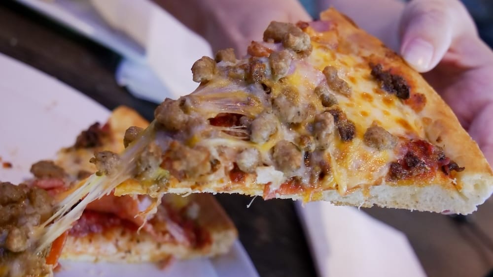 Primer plano de una porción de pizza Meatza en The Canadian Icehouse de Edmonton, Alberta, con trozos de carne dorada, queso derretido estirándose desde la pizza y un borde grueso sostenido con la mano.