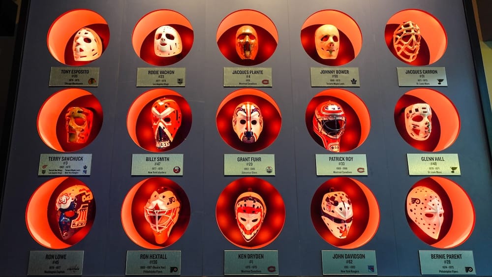 Exhibición iluminada de antiguas máscaras de arqueros de la NHL dentro de The Canadian Icehouse en Edmonton, Alberta, con placas de jugadores debajo de cada pieza de la colección del bar deportivo.