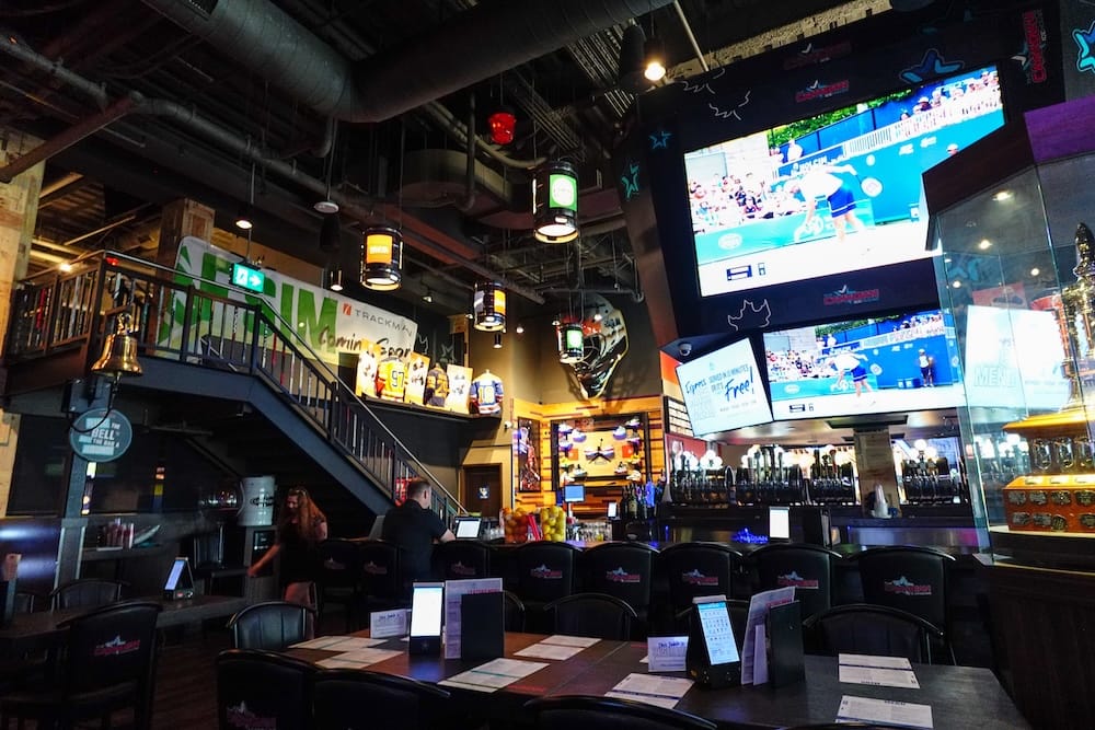 Interior de The Canadian Icehouse en Edmonton, Alberta, con grandes pantallas sobre la barra, recuerdos de la NHL, reservados y un diseño industrial propio de un bar deportivo.
