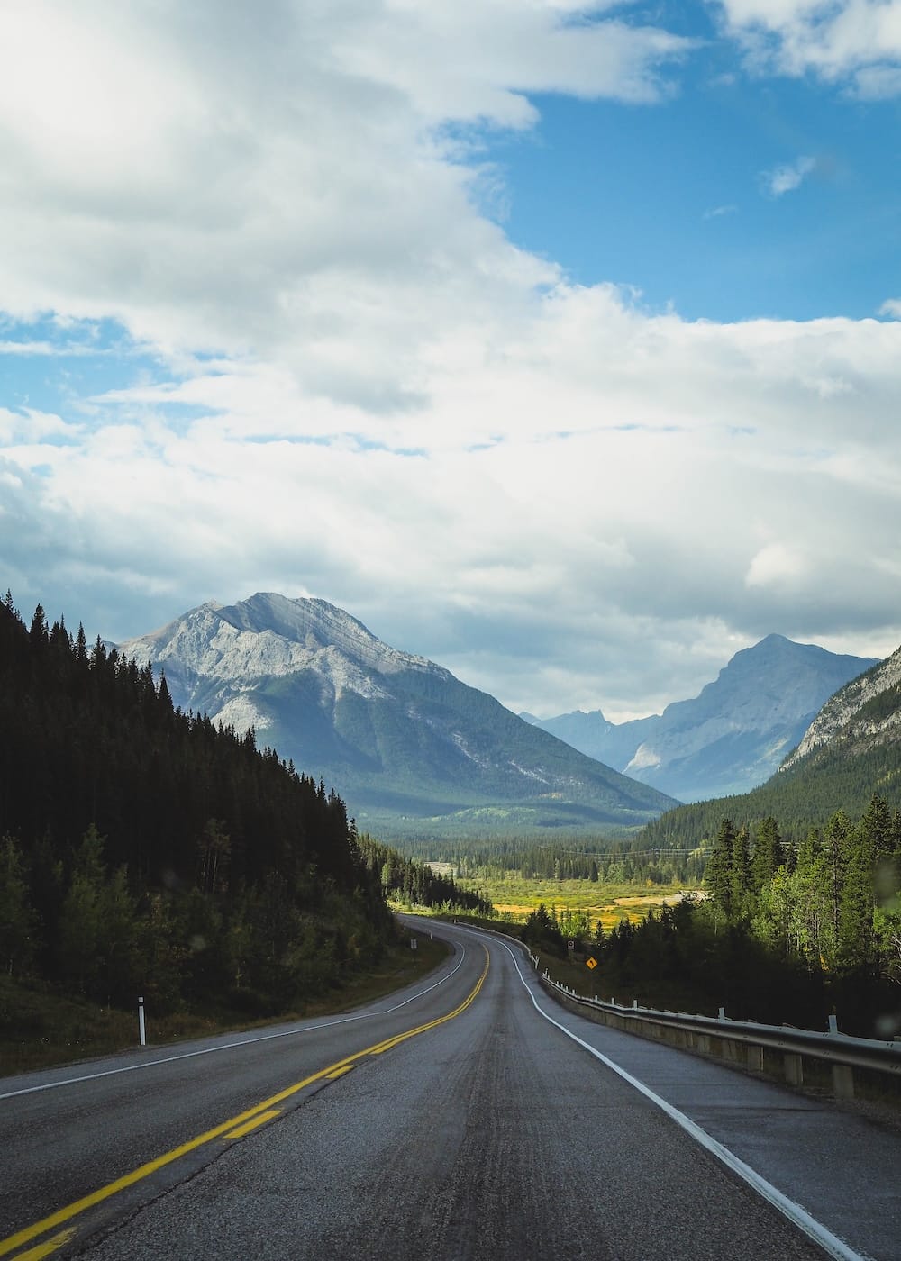 A two-lane scenic road curves through a forested valley in the Canadian Rockies, Alberta, Canada, with rugged mountain peaks ahead beneath large clouds and patches of blue sky.