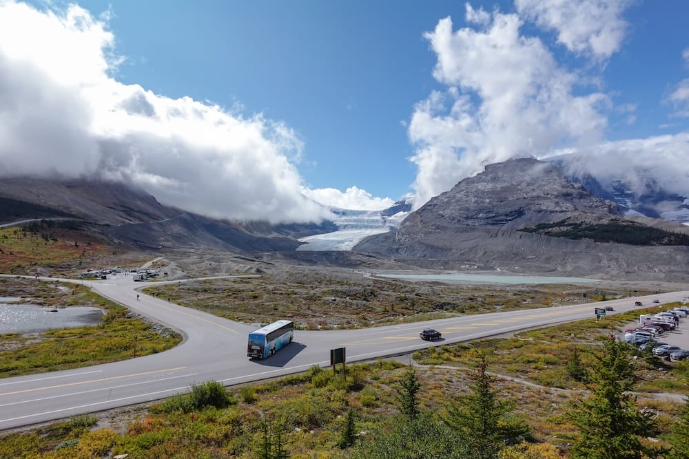 A tour bus and other vehicles travelling along a mountain road in the Canadian Rockies, Alberta, Canada, with a broad glacier, rocky slopes and low clouds filling the dramatic landscape behind them.
