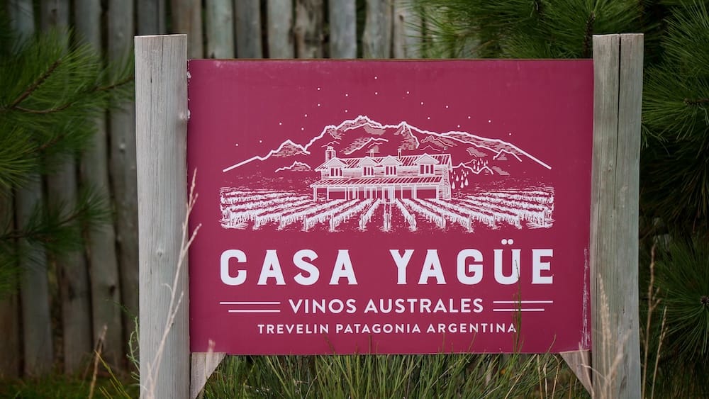 Casa Yagüe Vinos Australes vineyard sign in Trevelin, Chubut, Patagonia, Argentina A burgundy Casa Yagüe Vinos Australes sign stands beside pine trees in Trevelin, Chubut, Patagonia, Argentina, identifying the vineyard and winery with an illustration of vines, buildings and mountains.