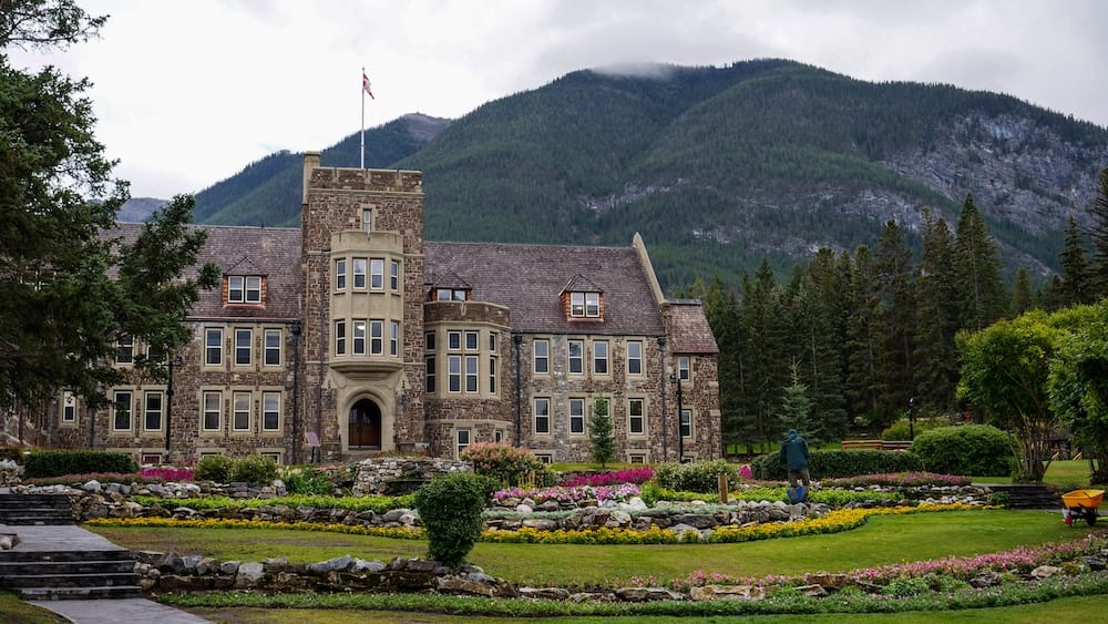 Wide view of the Banff Park Administration Building behind landscaped flower beds at Cascade of Time Garden in Banff, Alberta, Canada, with dense evergreen forest and a cloud-topped mountain rising behind the grounds.
