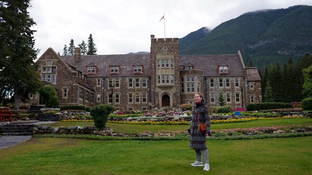 Audrey Bergner stands on the lawn at Cascade of Time Garden in Banff, Alberta, Canada, with the stone Banff Park Administration Building, flower beds and forested mountain slopes filling the background.