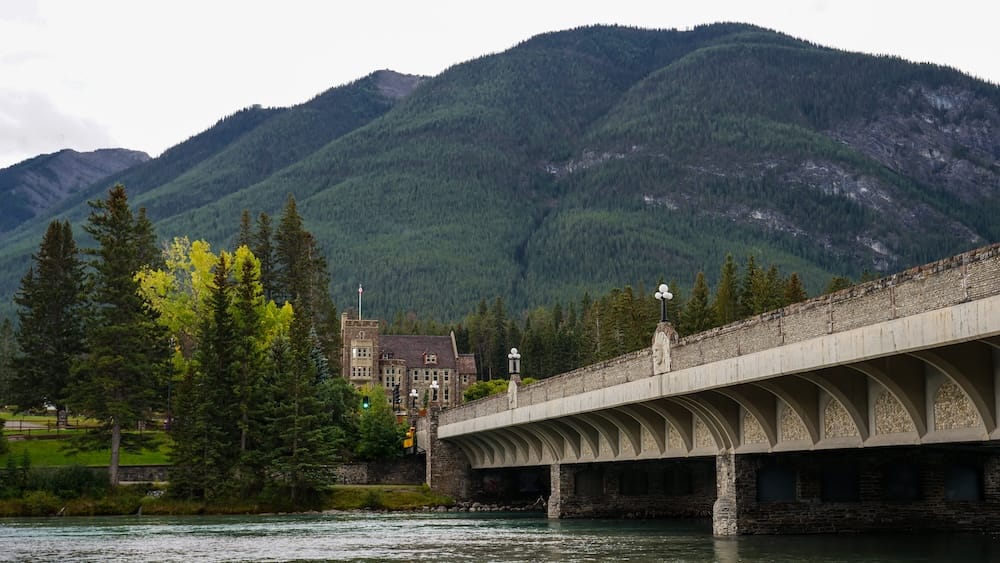 View across the Bow River bridge toward Cascade of Time Garden and the Banff Park Administration Building in Banff, Alberta, Canada, with dense evergreen forest and a mountain rising behind the town.