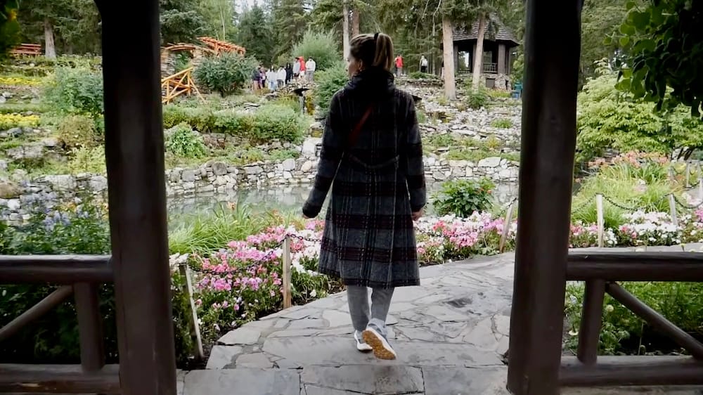 Audrey Bergner walks along a stone path through Cascade of Time Garden in Banff, Alberta, Canada, passing pink flower beds and a small pond with terraced landscaping and rustic garden structures ahead.