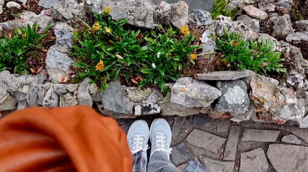 Downward creative view of Audrey Bergner’s white sneakers on a stone path beside a rock-edged flower bed at Cascade of Time Garden in Banff, Alberta, Canada, with small blooms and greenery filling the border.