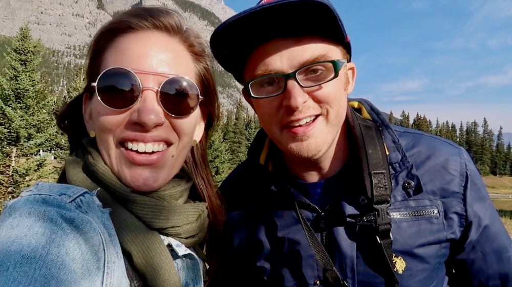 Audrey Bergner and Samuel Jeffery smile for a close selfie at Cascade Ponds in Banff National Park, Alberta, Canada, with evergreen forest, rocky mountain slopes and blue sky behind them.