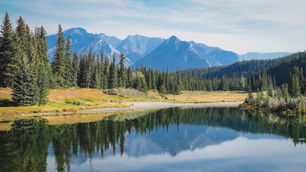 Cascade Ponds in Banff National Park, Alberta, Canada, with still reflective water, grassy picnic areas, evergreen forest and layered Rocky Mountain peaks rising in the background.