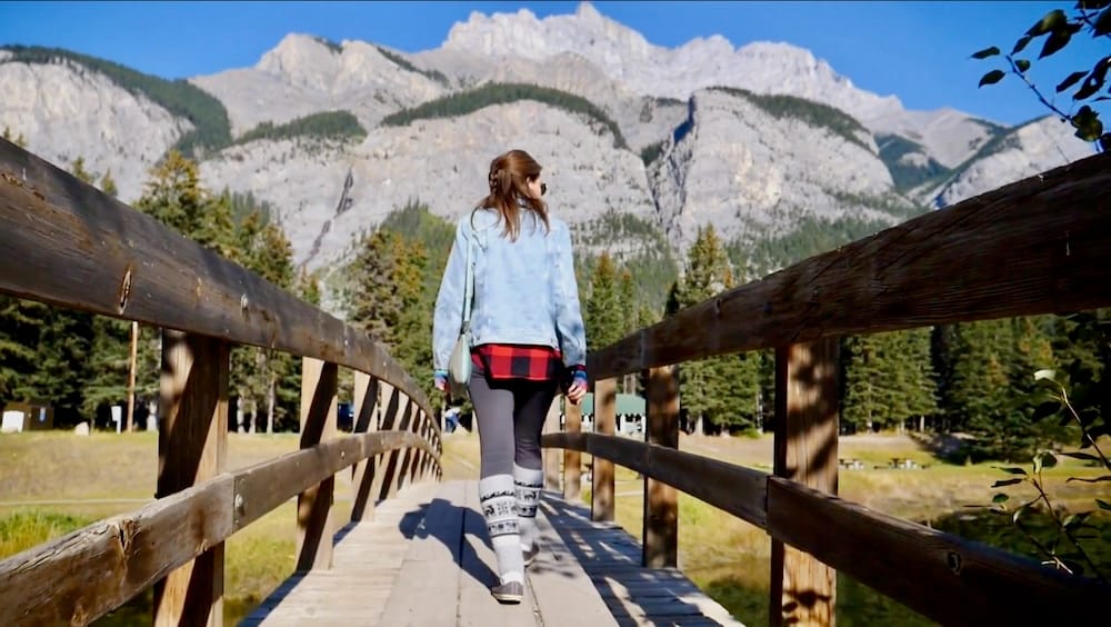 Cascade Ponds Banff National Park Alberta Canada Wooden Bridge Walk With Audrey Bergner and Mountain Scenery Audrey Bergner walking across a wooden bridge at Cascade Ponds in Banff National Park, Alberta, Canada, with evergreen forest and broad Rocky Mountain cliffs rising behind her.