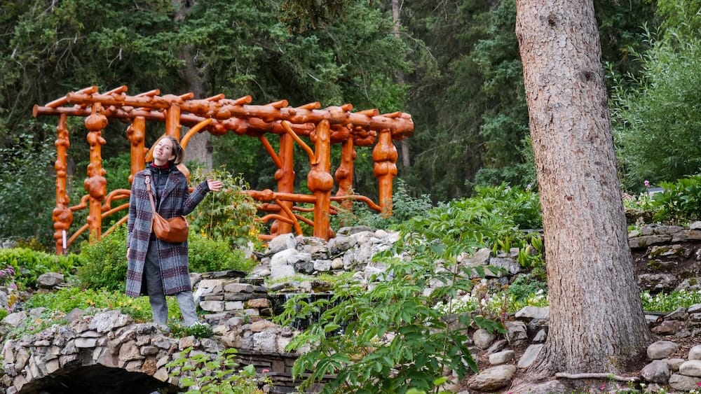 Audrey Bergner standing beside a rustic wooden garden structure and stone-lined water feature at Cascades of Time Garden in Banff, Alberta, Canada.