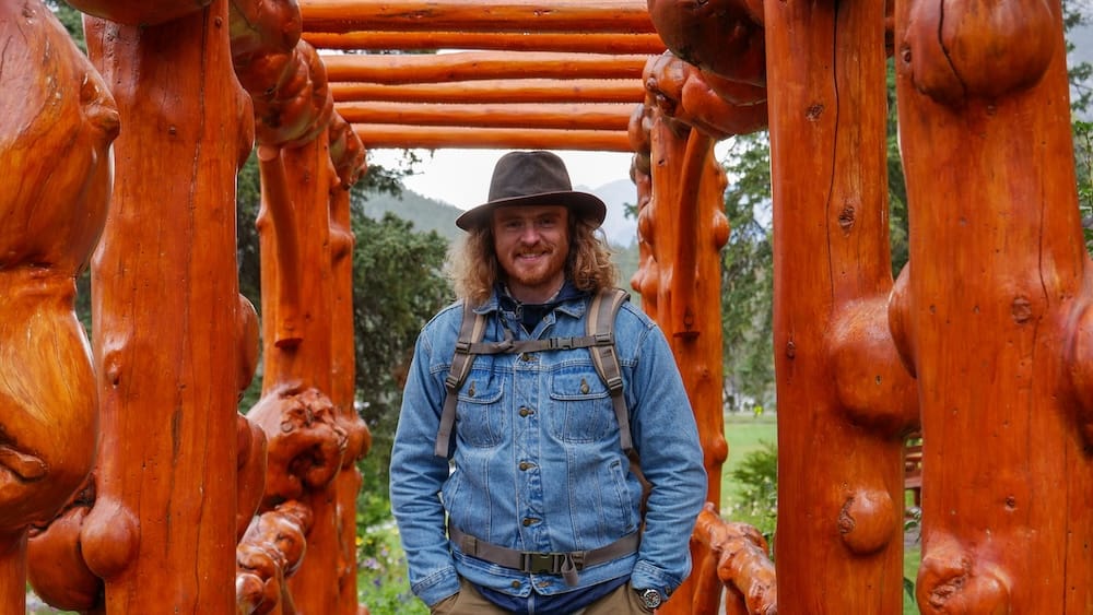Samuel Jeffery standing inside a rustic polished-log garden structure at Cascades of Time Garden in Banff, Alberta, Canada, with greenery and mountain scenery beyond.