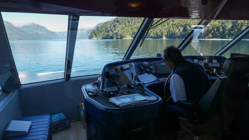 Un tripulante conduce un catamarán moderno por el lago Nahuel Huapi, con agua azul calma, costas boscosas y montañas lejanas visibles por las ventanas frontales cerca de Bariloche, Río Negro, Argentina.