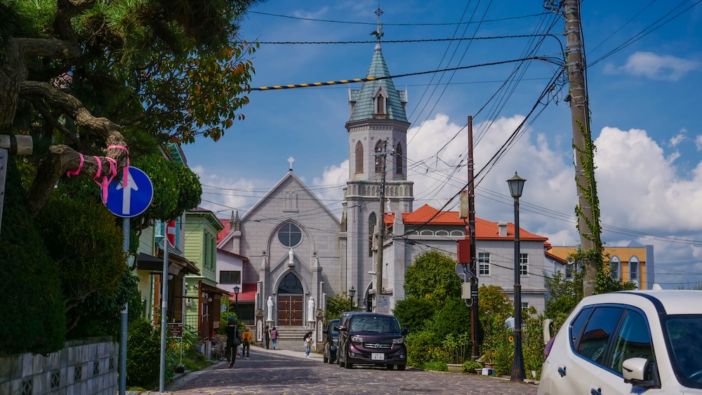 Catholic Motomachi Church at the end of a sloping street in Hakodate, Hokkaido, Japan, with its tall bell tower, red-roofed buildings, utility wires and pedestrians in the historic district.