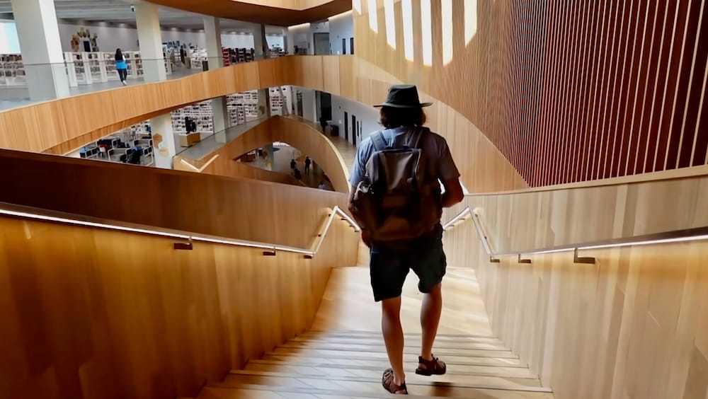 Samuel Jeffery descending a wood-lined staircase inside Calgary's Central Library, part of the city's free walkable downtown core, Alberta, Canada.