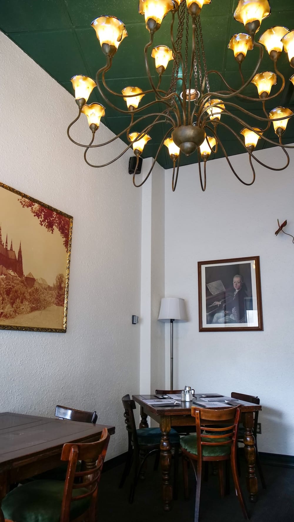 Una gran araña de bronce con pantallas de vidrio color ámbar cuelga de un techo verde en Bistro Praha en el centro de Edmonton, sobre un comedor con mesas de madera oscura, sillas tapizadas en verde y un retrato enmarcado en la pared.