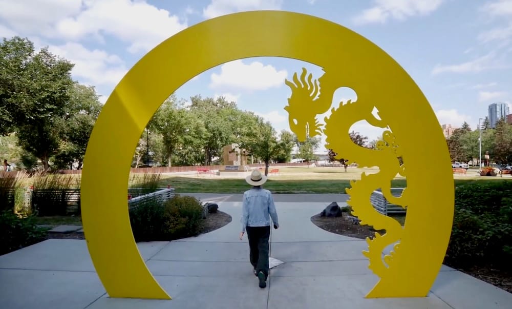 Audrey Bergner walks through a large yellow moon gate with a dragon cutout in Calgary’s Chinatown, with paved paths, trees and a few downtown buildings visible behind her in Calgary, Alberta, Canada.