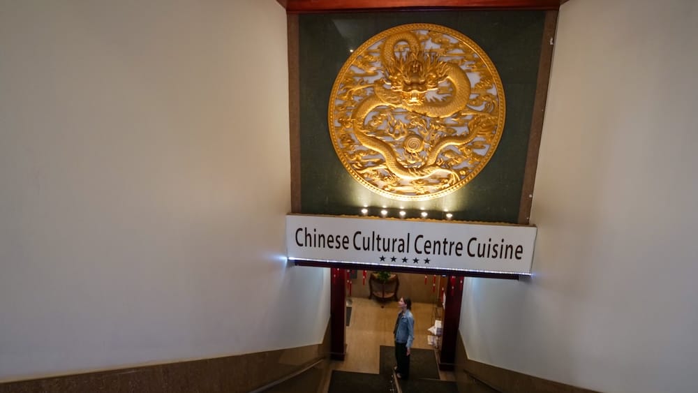 Audrey Bergner standing at the entrance to Chinese Cultural Centre Cuisine in Calgary Chinatown, Alberta, Canada, beneath the restaurant sign and a large golden dragon emblem.