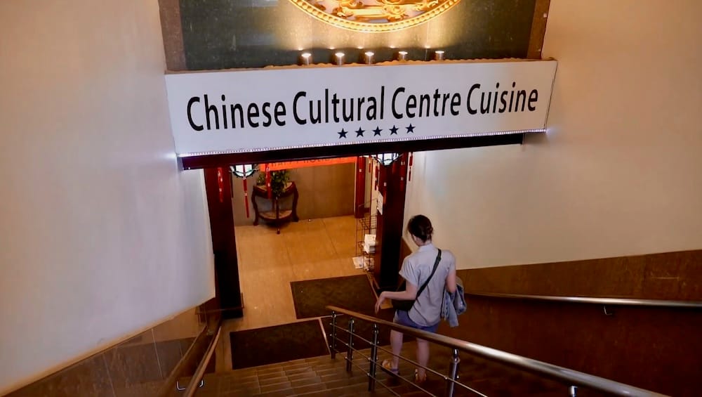Audrey Bergner descending a staircase toward the "Chinese Cultural Centre Cuisine" restaurant sign at the entrance within the Chinese Cultural Centre building in Calgary, Alberta, Canada.