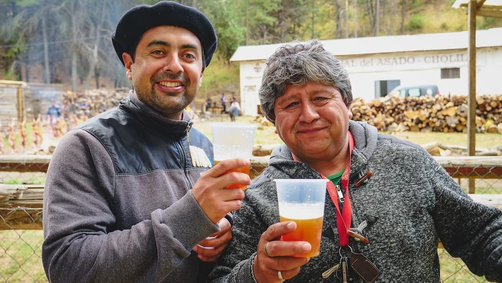 Two men toasting with plastic cups of golden craft beer at Fiesta Nacional del Asado in Cholila, Patagonia, Argentina, capturing the social, open-air drinking culture where beer, asado, and community gatherings come together in a relaxed rural setting.
