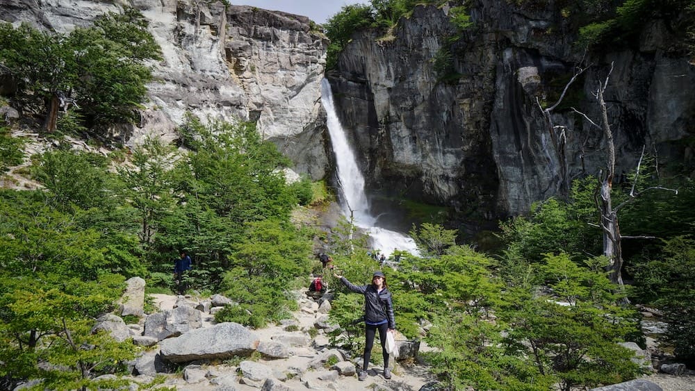 Chorrillo del Salto Waterfall Hike, El Chaltén, Santa Cruz Province, Argentine Patagonia — Audrey Bergner Audrey Bergner hiking toward Chorrillo del Salto waterfall near El Chaltén, Santa Cruz Province, in Argentine Patagonia, pausing on a rocky forest trail with other hikers visible below the cascading falls and granite cliffs.