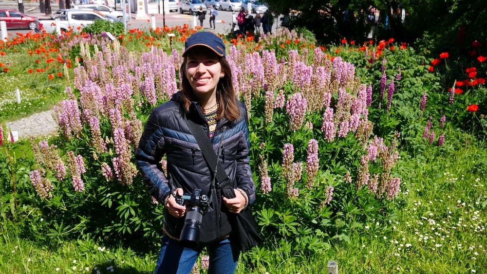 Audrey Bergner stands with a camera among tall pink and purple lupines in central Ushuaia, Tierra del Fuego, Patagonia, Argentina, with red flowers, parked cars and pedestrians visible behind the garden.