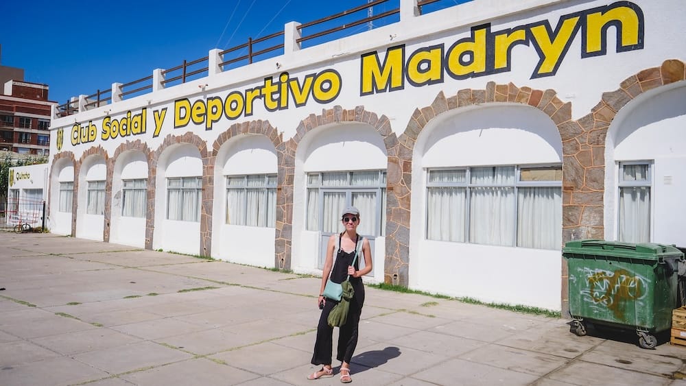 Audrey Bergner stands outside the white arched facade of Club Social y Deportivo Madryn in Puerto Madryn, Chubut, Patagonia, Argentina, beneath bright yellow signage on a sunny city street.