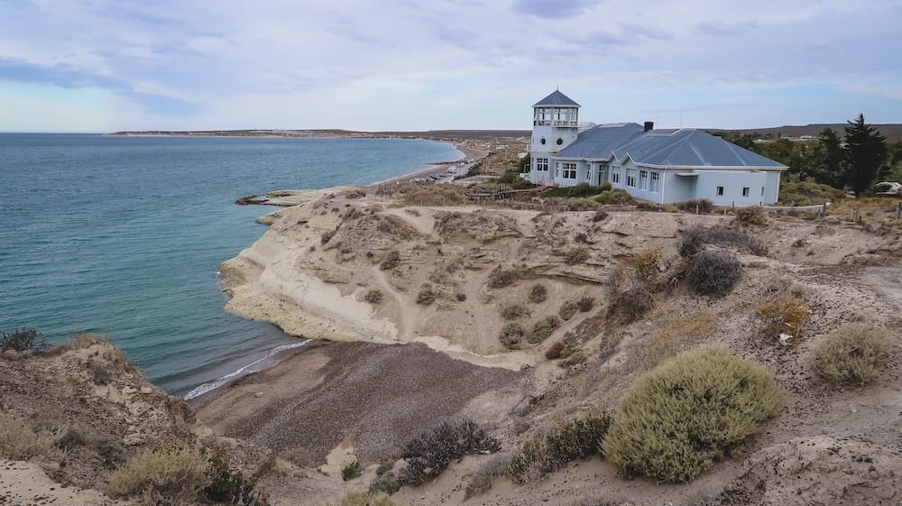 Coastal lookout building perched above dramatic eroded cliffs in Puerto Madryn, Argentina, with turquoise water below, a small cove at the base of the bluffs, and the rugged Patagonian shoreline stretching into the distance.
