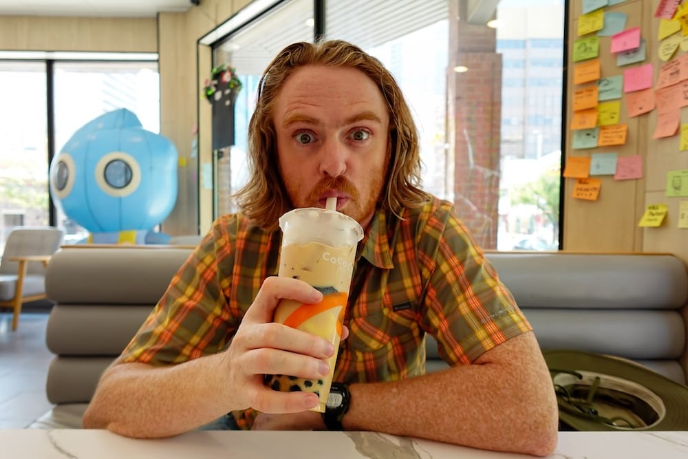 Samuel Jeffery tomando bubble tea en CoCo, Chinatown de Calgary, Alberta, Canadá Samuel Jeffery toma pearl milk tea dentro de CoCo en Chinatown de Calgary, Alberta, Canadá, sosteniendo un vaso de bubble tea con perlas de tapioca mientras está sentado junto a grandes ventanales que dan a la calle.