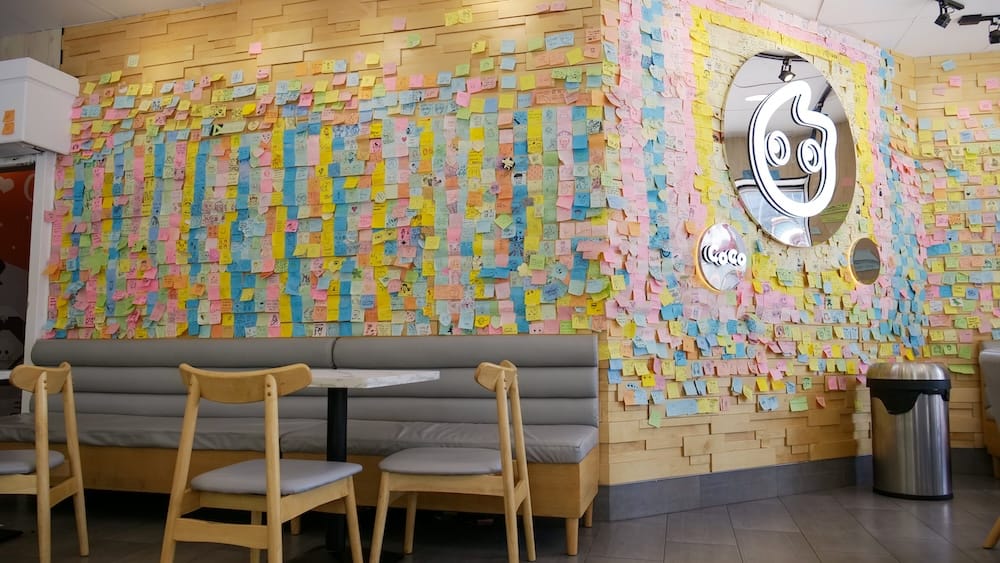 Booth seating beneath a wall almost entirely covered in colorful customer sticky notes at CoCo Fresh Tea & Juice in Chinatown, Calgary, Alberta, Canada, with the shop's lit mascot logo mounted in the center.