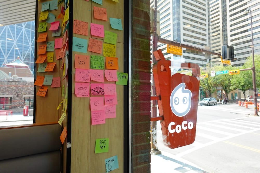CoCo Fresh Tea & Juice's cup-shaped storefront sign viewed from inside the shop, next to a wall covered in customer sticky notes, with Centre Street and downtown Calgary, Alberta, Canada, visible through the window.