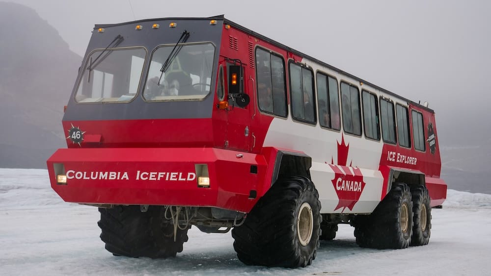 A red Ice Explorer vehicle parked on glacial ice at the Columbia Icefield in Alberta, Canada, with oversized tires, misty mountain surroundings and the snowy surface of the Canadian Rockies glacier experience.