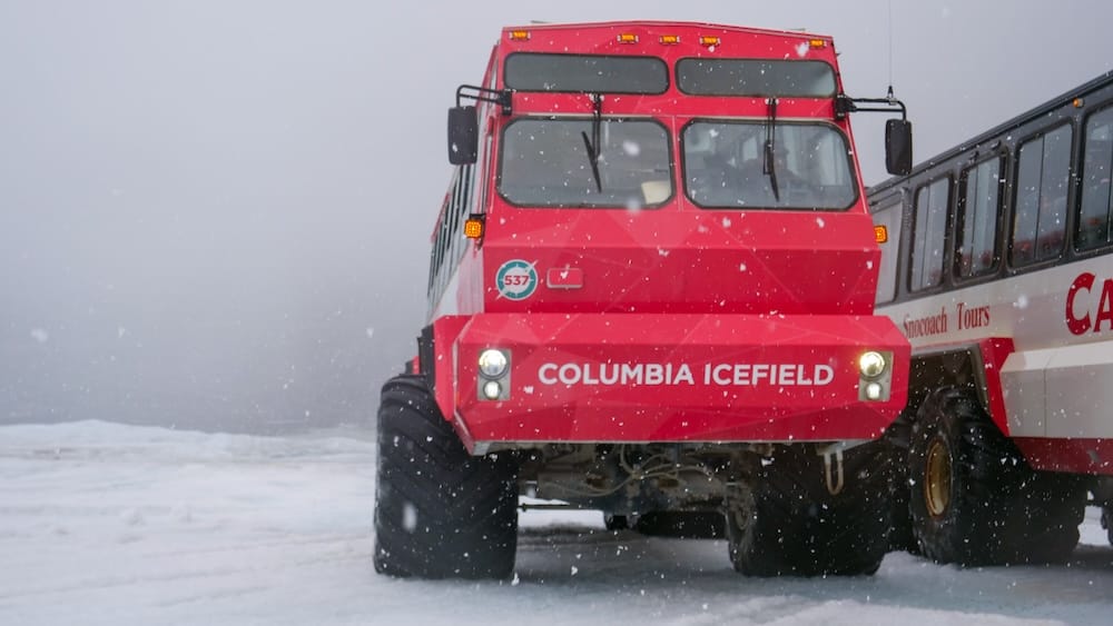A red Ice Explorer vehicle on the snowy Columbia Icefield in Alberta, Canada, with oversized tires, falling snow and another glacier tour vehicle parked beside it in the Canadian Rockies.