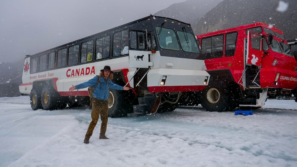 Samuel Jeffery en el Columbia Icefield, Alberta, Canadá, junto a vehículos Ice Explorer bajo una nevada Samuel Jeffery bajo una nevada junto a grandes vehículos Ice Explorer en el Columbia Icefield, Alberta, Canadá, con el suelo cubierto de nieve y laderas oscuras de montaña detrás.