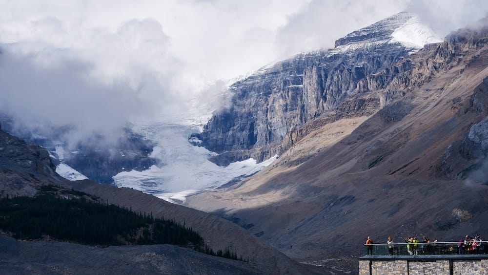 Visitantes observan el paisaje desde la terraza exterior del Columbia Icefield Discovery Centre, Alberta, Canadá, con terreno glaciar, roca expuesta y nubes bajas sobre las montañas.