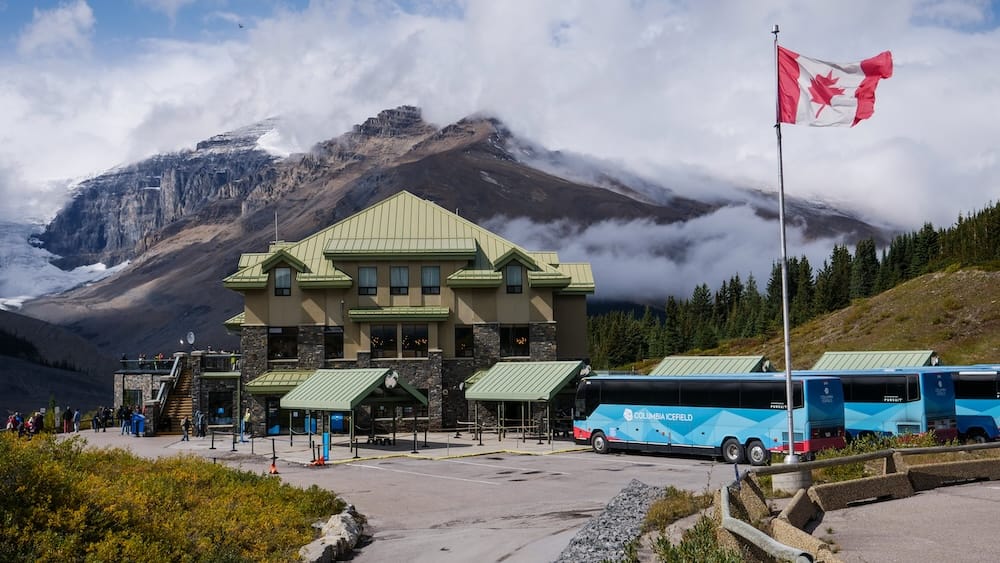 Columbia Icefield Discovery Centre en el Parque Nacional Jasper, Alberta, Canadá, con micros estacionados afuera, una bandera canadiense junto al edificio y montañas con glaciares detrás.