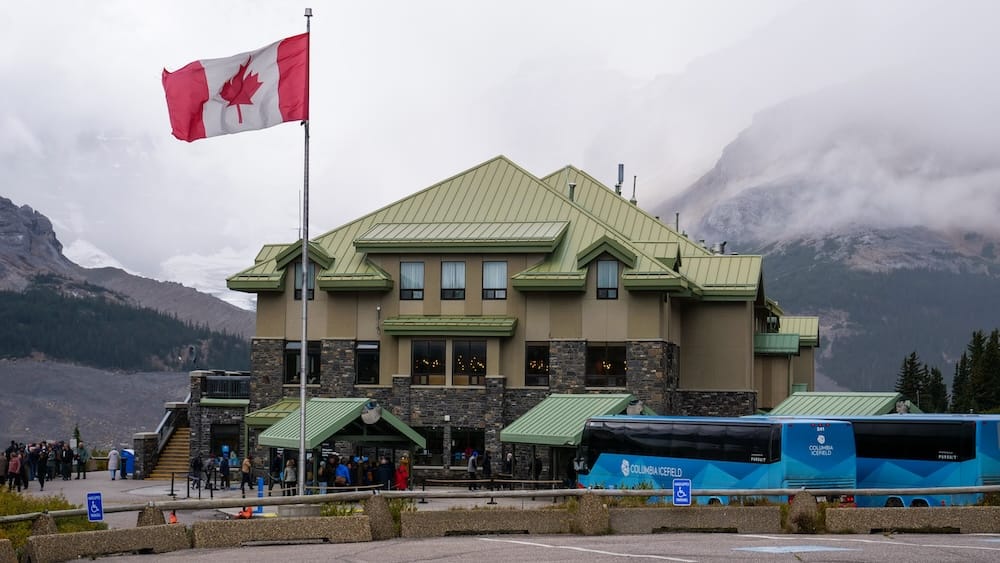 El edificio del Columbia Icefield Discovery Centre con una bandera canadiense flameando en el frente y un micro de excursión al Columbia Icefield estacionado en la entrada, montañas y niebla detrás, Alberta, Canadá.