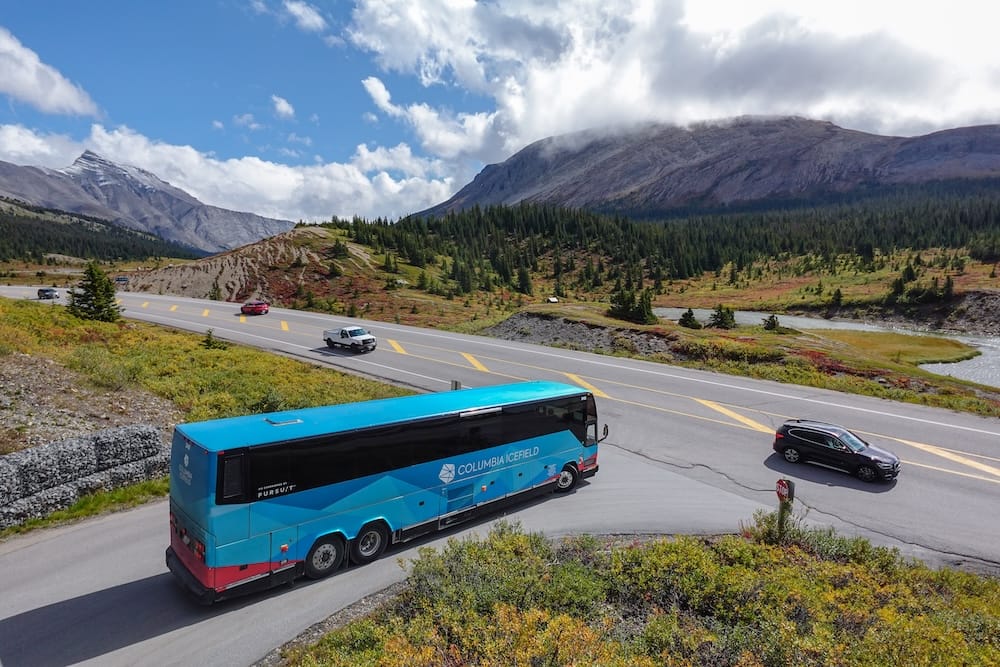 Un micro de excursión con la marca del Columbia Icefield y varios vehículos personales compartiendo la ruta Icefields Parkway, con montañas y un pequeño lago visibles al costado del camino, Alberta, Canadá.