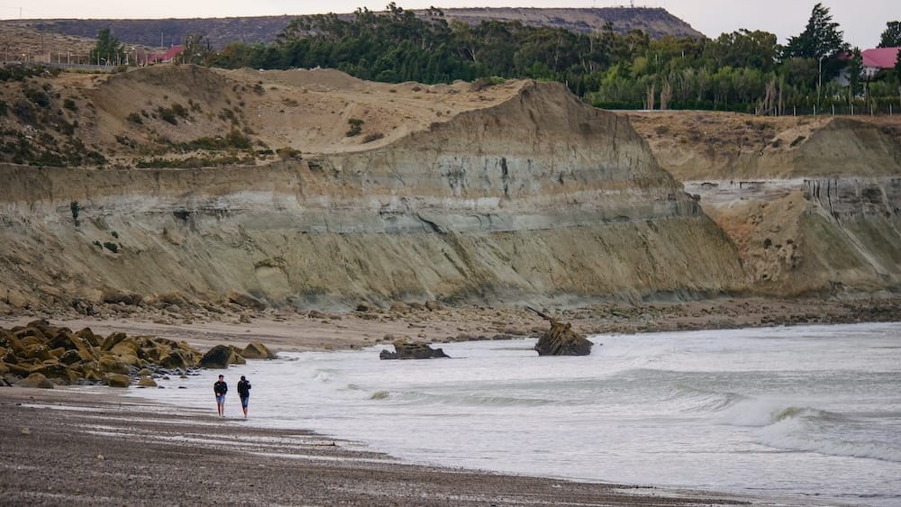 Comodoro Rivadavia Patagonia coastal cliffs with travelers walking along rugged Atlantic shoreline in Argentina Two travelers walking along the rugged eroded coastal cliffs of Comodoro Rivadavia Chubut Argentina with waves breaking along the remote Patagonian Atlantic shoreline under a muted coastal sky.