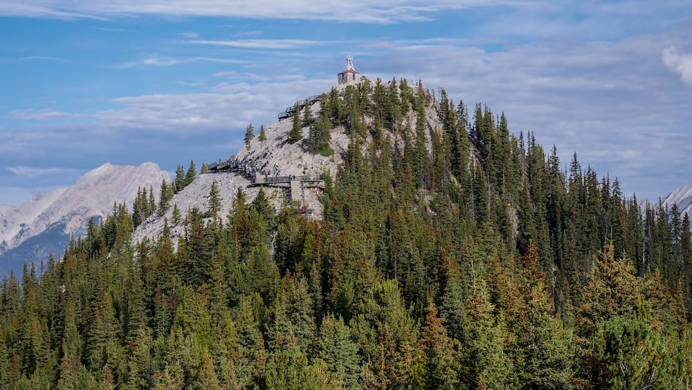 Cosmic Ray Station atop Sanson's Peak from the Banff Gondola summit, Sulphur Mountain, Alberta, Canada The Cosmic Ray Station and weather tower sit atop Sanson's Peak, reached by a boardwalk visible switchbacking up the ridge, with forested slopes and mountains behind on Sulphur Mountain, Banff, Alberta, Canada.