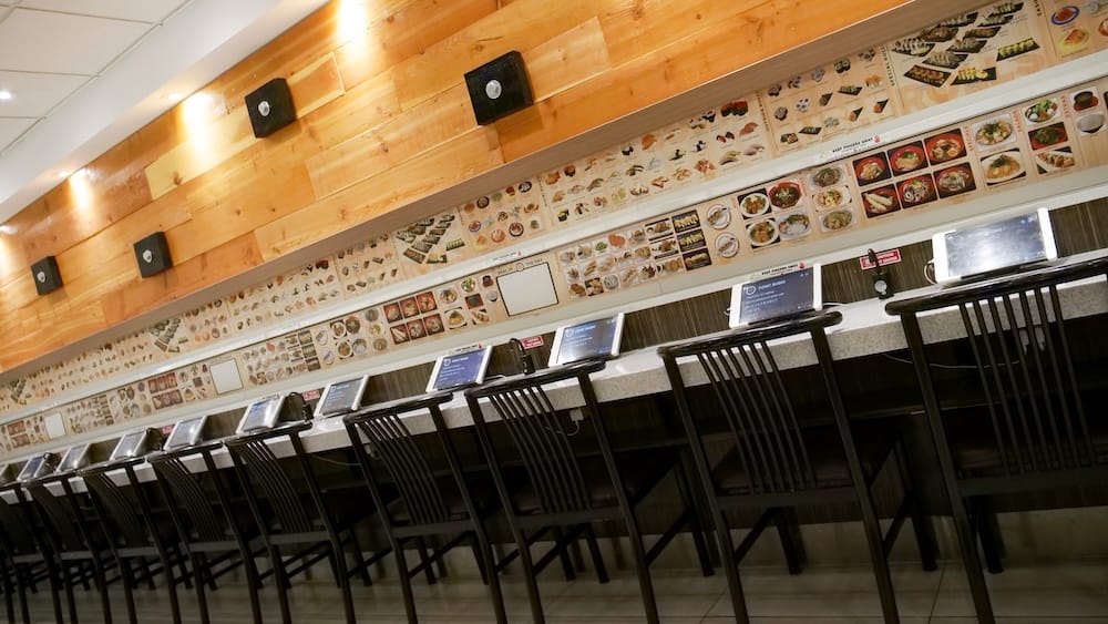 Fila de asientos en la barra, cada uno con su propia tablet para pedir, debajo de una pared con fotos ilustradas del menú, en Point Sushi en Calgary, Alberta, Canadá.