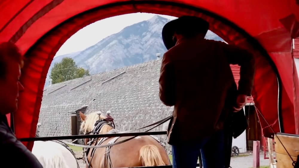 Salida de carruaje cubierto, Banff Trail Riders, Banff, Alberta, Canadá Pasajeros dentro de un carruaje cubierto rojo miran al conductor y los caballos partir junto a un granero de madera en Banff, Alberta, Canadá, al inicio del paseo Wagon Cowboy Cookout.