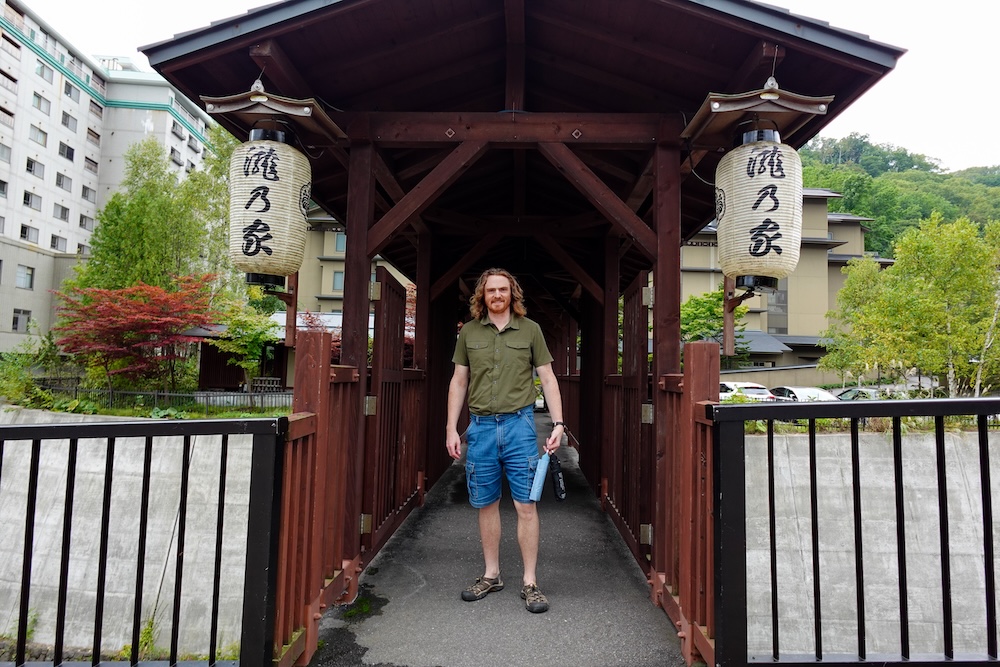 Samuel Jeffery stands at the entrance to a covered wooden footbridge hung with two paper lanterns. Large hotels and green, forested hills rise behind him in Noboribetsu Onsen, Hokkaido, Japan.